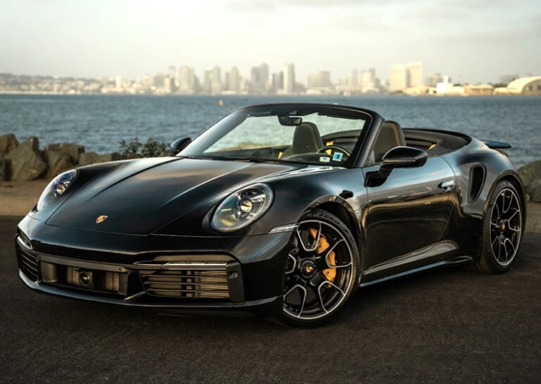 Black convertible sports car parked near a body of water with a city skyline in the background